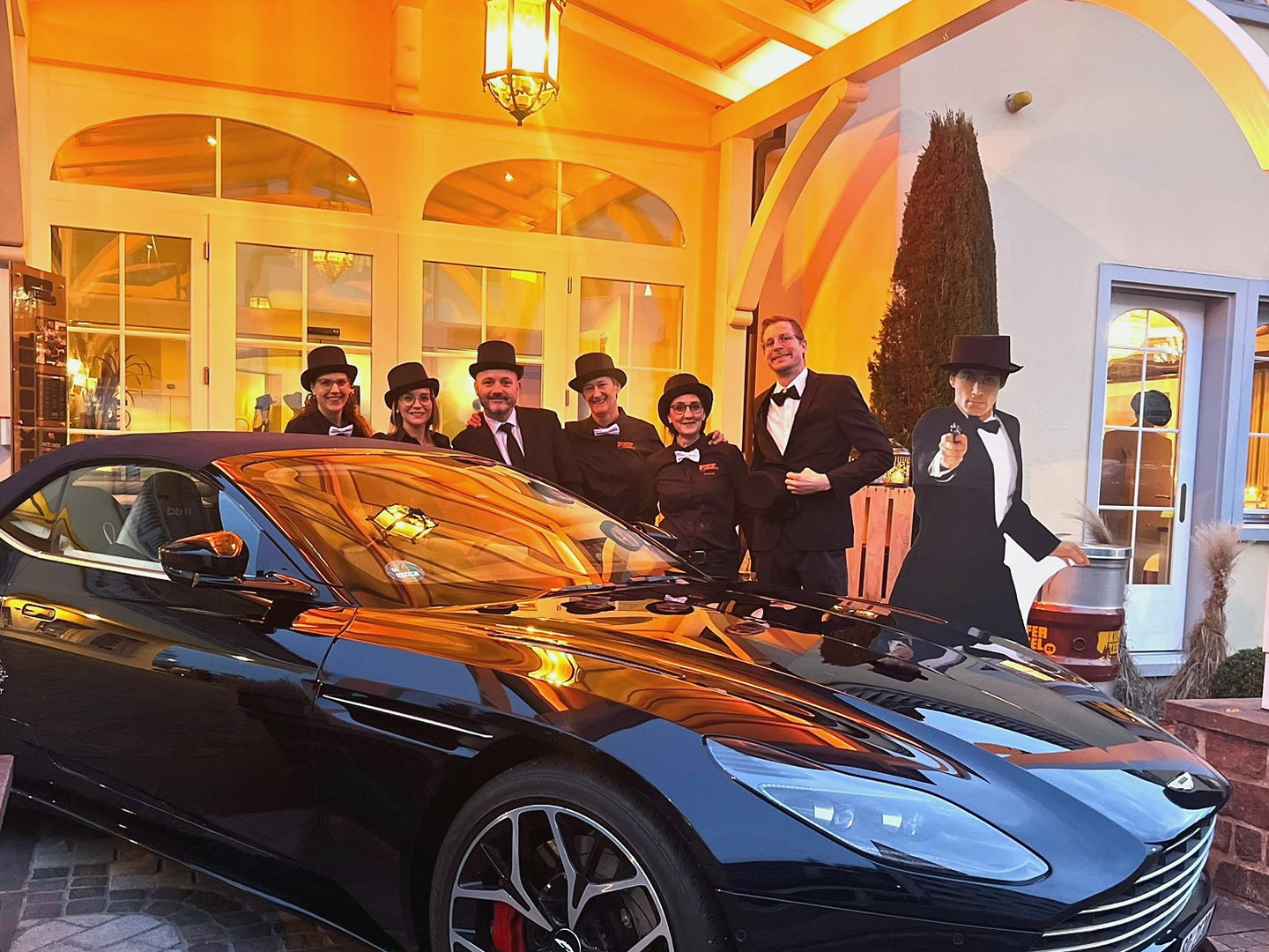 Team in Zylindern und Fliegen neben einem schwarzen Aston Martin an einem Motto-Abend